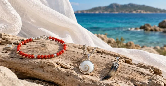 Sélection de bijoux corses en corail rouge, nacre, corne et argent posés sur du bois flotté devant une mer turquoise et un drapé blanc.