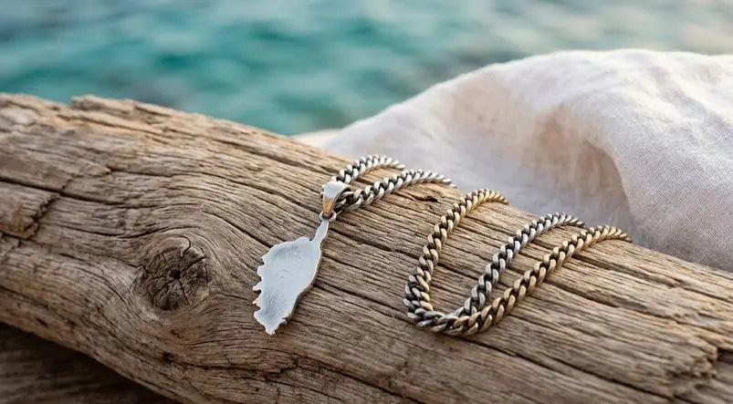 Pendentif en forme de Corse avec sa chaîne, posé sur du bois flotté devant une mer turquoise et un drapé blanc.
