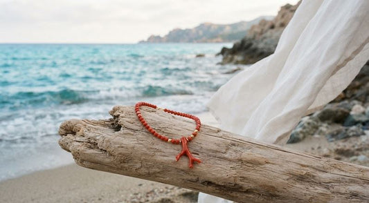 Bijou artisanal en corail rouge posé sur du bois flotté devant une mer turquoise et un drapé blanc