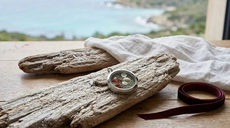 Pendentif rond posé sur du bois flotté, avec un ruban bordeaux et un drapé blanc devant la mer.