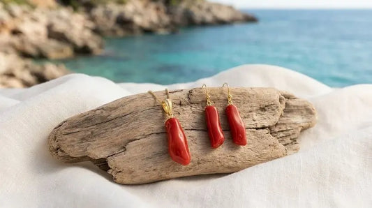 Pendentif et boucles d’oreilles en corail rouge aux attaches dorées, sur du bois flotté et un drapé blanc devant une mer turquoise.